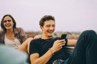 Cheerful man looking at smart phone while female friend having fun at rooftop party