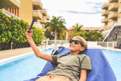 Smiling boy taking selfie while lying against swimming pool
