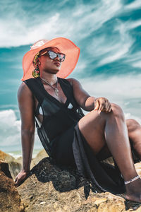 African woman with sunglasses and sun hat sitting on a rock by the tropical beach in ghana