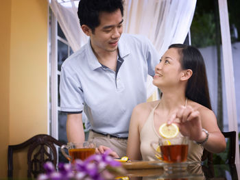 Smiling woman with man adding lemon slice in tea on glass table at home