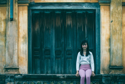 Portrait of young woman sitting on retaining wall against door