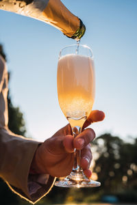 Crop anonymous person having champagne poured into glass from bottle on blurred background of outdoor poolside in evening