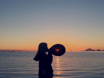 Side view of woman blowing balloon at sea shore against sky during sunset