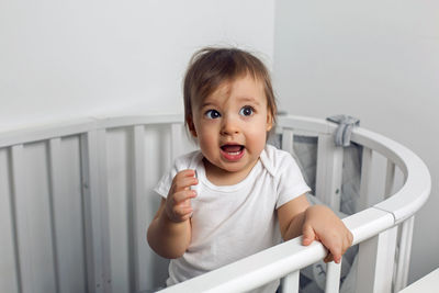 One year old child in white clothes standing in a white round bed in his nursery