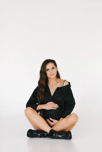 A stylish pregnant woman in a black shirt and boots sits on a white background and hugs her stomach
