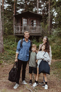 Full length portrait of family with luggage standing against house in forest