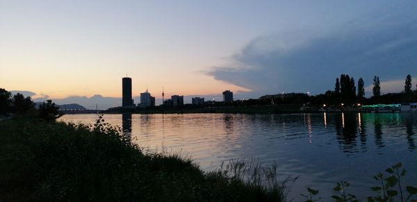 Silhouette buildings by river against sky at sunset