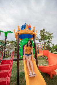 Young woman sitting on slide at playground against sky