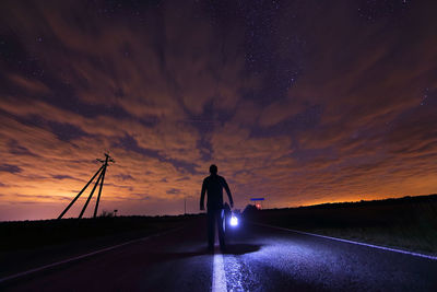 Silhouette man carrying torch light standing on road against cloudy sky