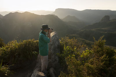 Man photographing mountain while standing on rock against sky