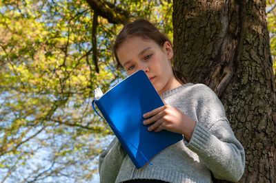 Young school age girl reading from blue textbook in park leaning against tree.