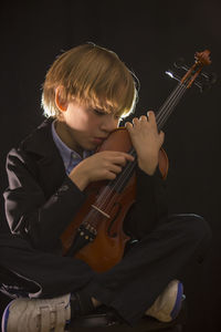 Boy holding violin while sitting against black background