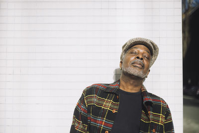 Mature man wearing flat cap while standing with eyes closed against wall