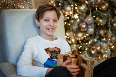 Happy boy sitting by the christmas tree with a gift