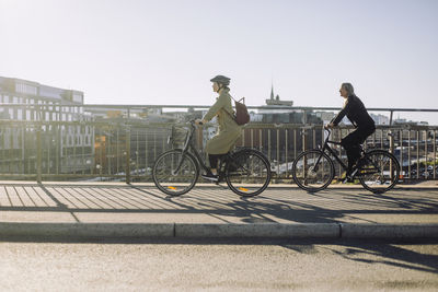 Side view of female business professionals riding bicycle on lane during sunny day