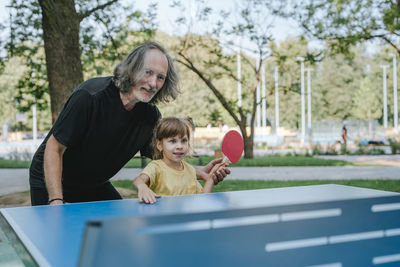 Happy grandfather and granddaughter playing table tennis in park