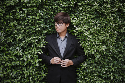 A young asian man was wearing a suit in front of a tree bush and looking sideways.