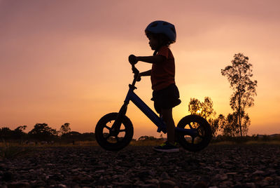 Silhouette boy riding bicycle on field against sky during sunset