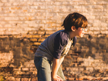Side view of boy standing against brick wall on sunny day