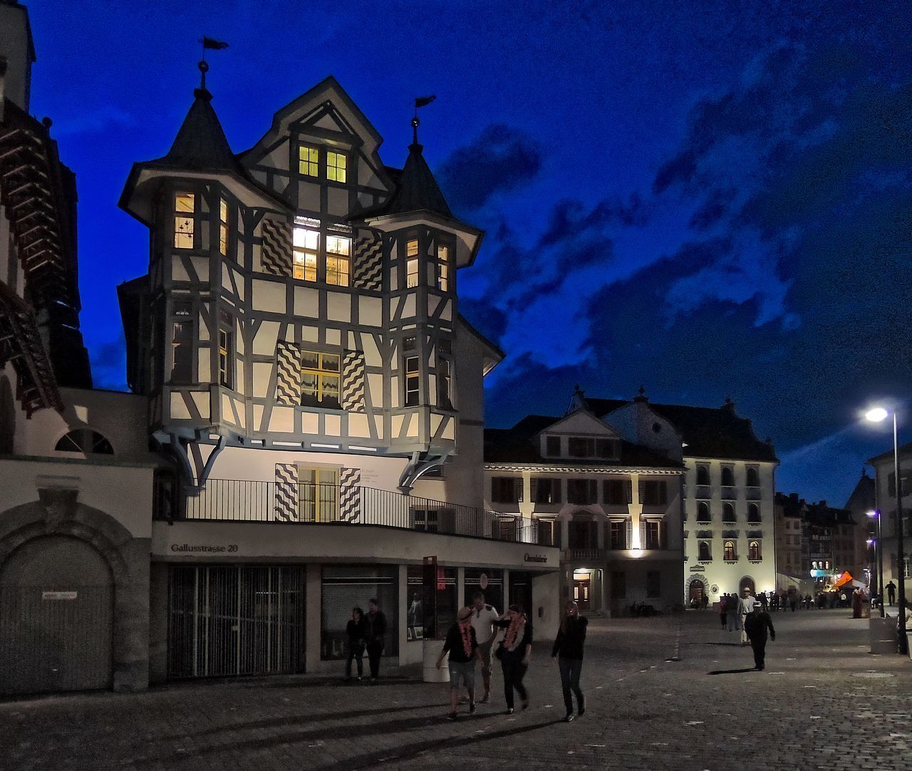 PEOPLE WALKING ON ILLUMINATED STREET AMIDST BUILDINGS AT NIGHT