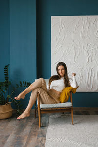 Beautiful young woman in beige trousers and a jumper is sitting at home in an armchair and dreaming