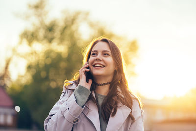 Smiling beautiful girl with long hair in a grey trench coat using smartphone in the spring 