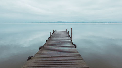 Narrow wooden jetty leading to calm sea against cloudy sky