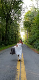 Woman holding suitcase while running on road amidst trees