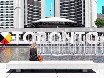 Woman sitting on fountain against building in city