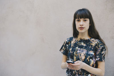 Portrait of young woman holding smart phone while standing against wall