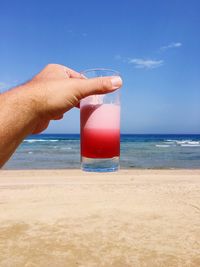 Close-up of hand holding juice in glass at beach against sky