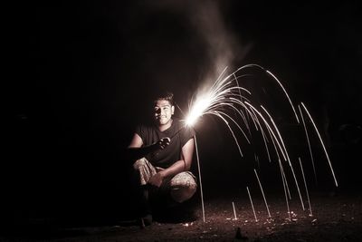 Portrait of mature man holding firework while crouching on field at night