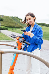 Content female renting parked electric scooter in city and browsing mobile phone