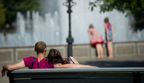 Rear view of man and woman sitting on bench during sunny day