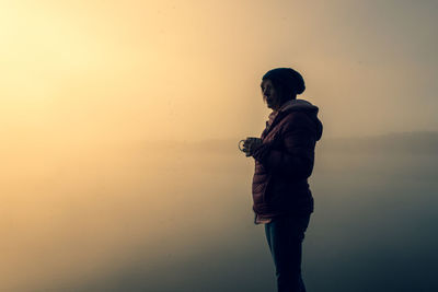 Side view of woman standing by lake against sky during dawn