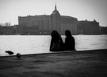 Silhouette friends sitting by river in city