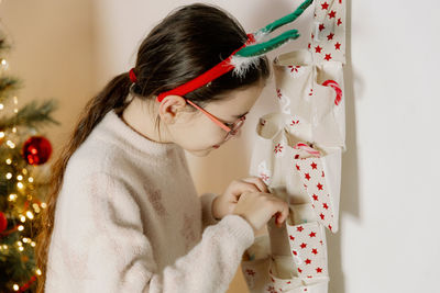 A girl takes out a gift from an advent calendar while standing near the christmas tree.