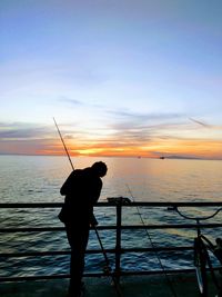 Silhouette man adjusting fishing rods on railing in sea against sky