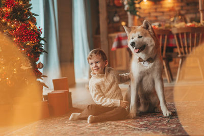 Candid authentic happy little boy in knitted beige sweater hugs dog with bow tie at home on xmas