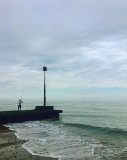 Rear view of man fishing at beach against cloudy sky