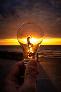 A man holds up a lightbulb in front of a woman's ballerina silhouette on the beach at sunrise 