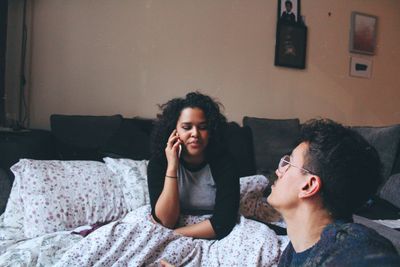 Young man looking at woman talking on smart phone while sitting on bed at home