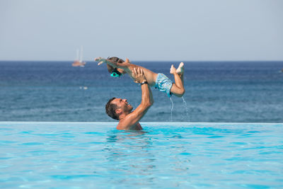 Side view of father holding son in mid air against sea