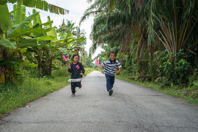 Full length of girl with brother holding malaysian flags on road