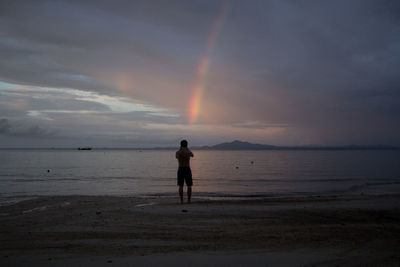 Rear view of man standing on beach against sky during sunset