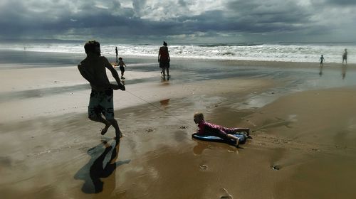 Man pulling daughter on surfboard at beach against cloudy sky