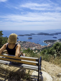 Rear view of woman looking at sea while sitting on bench against blue sky