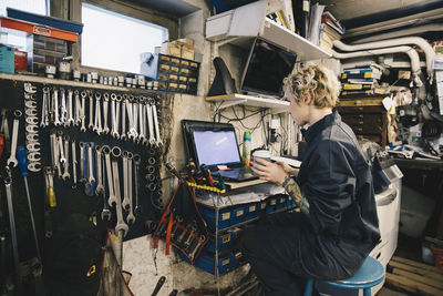 Side view of female mechanic holding disposable cup while using laptop at auto repair shop