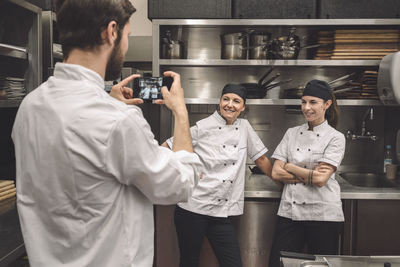 Male chef taking photograph of smiling female coworkers in commercial kitchen