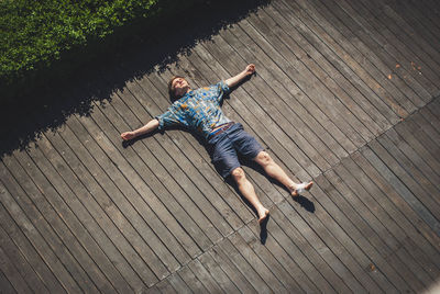 High angle view of carefree man with arms outstretched lying on boardwalk